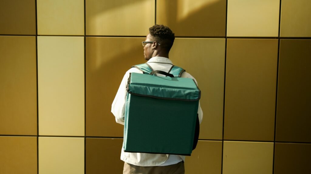 pexels-photo-8989328-8989328 Back view of a delivery person standing against a golden tiled wall with a large backpack.