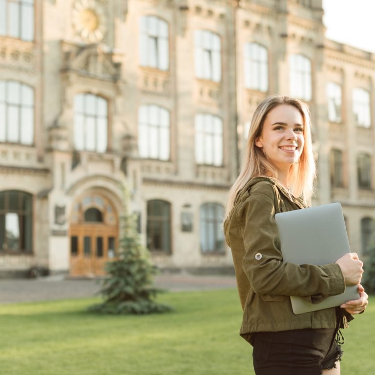 Portrait of happy lady in casual clothes standing on grass in campus on background of university building with laptop in hands, looking away at copy space and smiling.Girl is walking college yard